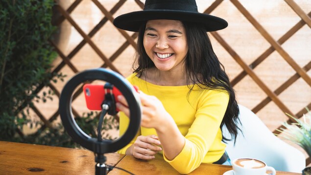Une jeune femme sourit vers son téléphone, assise à une table sur laquelle se trouve un cercle lumineux, et un café.