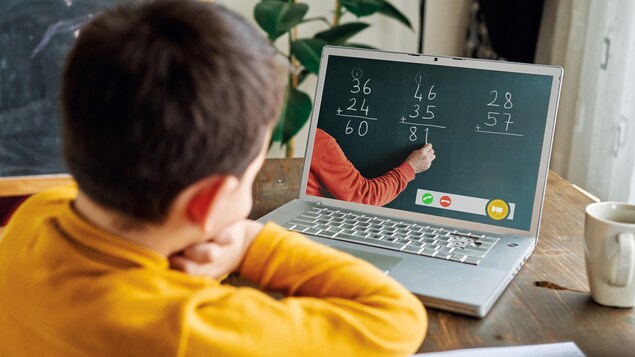 Un enfant devant un ordinateur regarde son enseignant écrire des additions au tableau.