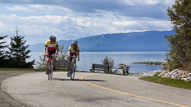Des cyclistes roulent sur une petite route de l'Isle-aux-Coudres. Le fleuve et les montagnes forment l'arrière-plan.