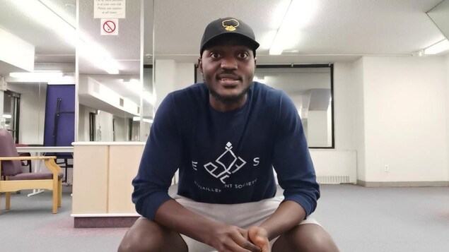 Un black trentenaire habillé en sportif avec une casquette assis dans un studio