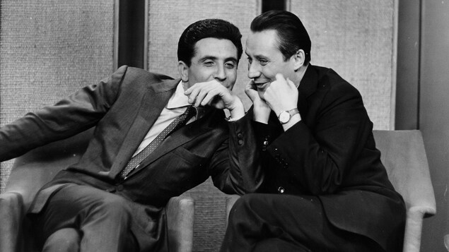 Jacques Normand semble chuchoter à l'oreille de Gilbert Bécaud alors qu'ils sont assis côte à côte. La photographie a été prise dans le studio de l'émission Les Couche-Tard, le 15 février 1961. Jacques Normand semble chuchoter à l'oreille de Gilbert Bécaud alors qu'ils sont assis côte à côte. La photographie a été prise dans le studio de l'émission Les Couche-Tard, le 15 février 1961.