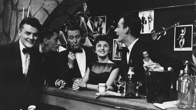 L'animateur Jacques Normand fumant la pipe et debout derrière un bar est accompagné par Roger Baulu et par les comédiens Dominique Michel et Pierre Thériault. L'animateur Jacques Normand fumant la pipe et debout derrière un bar est accompagné par Roger Baulu et par les comédiens Dominique Michel et Pierre Thériault.