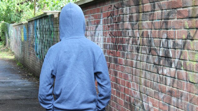 Un adolescent de dos portant un chandail à capuchon marche le long d'un mur de briques. 
