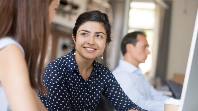 Une jeune femme sourit et discute avec une collègue dans un espace de travail.