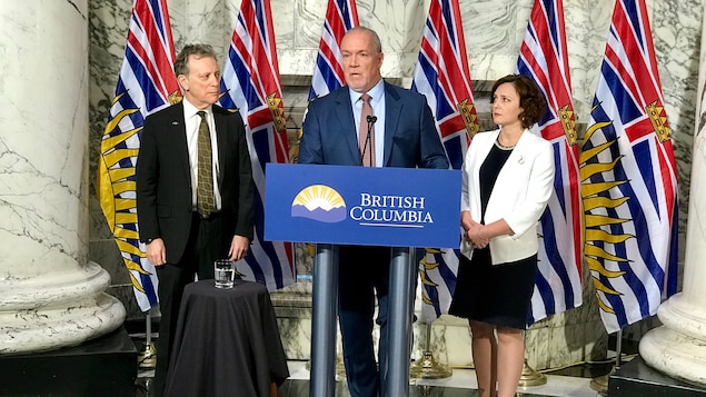 Le premier ministre de la Colombie-Britannique John Horgan est au podium avec les ministres George Heyman et Michelle Mungall à ses côtés.
