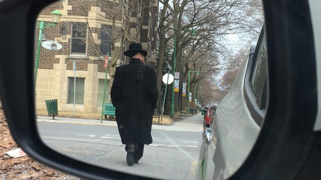 Gros plan du rétroviseur d'une voiture dans lequel on voit un homme de dos habillé de noir et portant un chapeau noir, en train de marcher dans la rue.