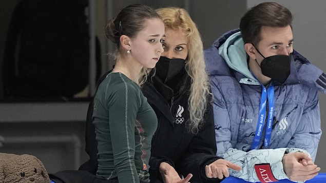 La jeune patineuse regarde la glace d'un air inquiet, pendant que son entraîneuse lui parle, accotée sur la bande de la patinoire. Un homme du Comité olympique russe, non identifié sur la photo, se trouve à leur gauche. 