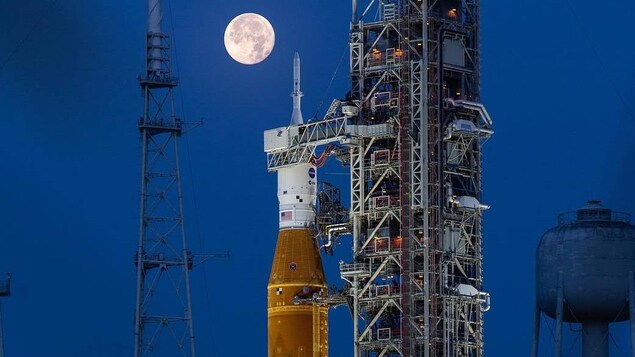 Vue du lanceur SLS sur son pas de tir avec la Lune en arrière plan.