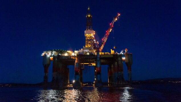 Une plateforme pétrolière éclairée, au large des côtes, la nuit, illumine la surface de l'eau. 