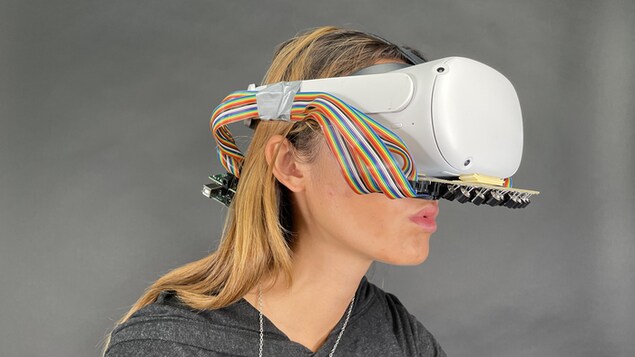 Une jeune femme aux cheveux long porte un casque de réalité virtuelle, qui ressemble à des grosses lunettes de ski. Sous le dispositif se trouve une série de haut-parleurs miniatures.