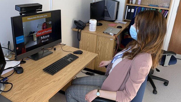 Une jeune femme à la longue chevelure brune, portant un masque, est assise devant un écran d'ordinateur qui est équipé de divers instruments vidéo.
