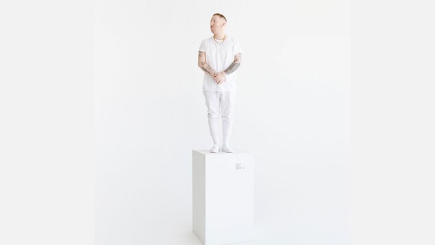 Un homme tout en blanc se tient sur un socle blanc dans un décor immaculé. 
