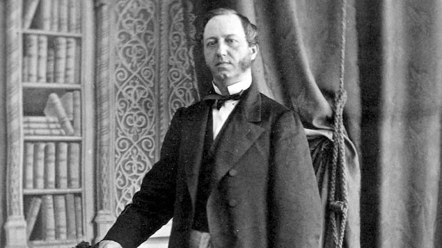 L'honorable Pierre-Joseph-Olivier Chauveau, debout devant une chaise, chapeau haut-de-forme à la main.