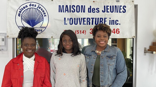 Deux employées et une habituée de la maison des jeunes L'Ouverture se tiennent devant une banderole à l'effigie de l'établissement.