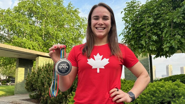 Linda Morais de Tecumseh, souriante, portant un chandail avec une feuille d'érable, est debout devant des arbustes et tient dans sa main une médaille de bronze sur laquelle est inscrite Paris 2024 Qualifier.