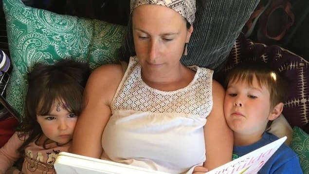 Deux enfants sont dans un lit, un de chaque côté de leur mère et lisent un livre que la femme tient dans ses mains. 