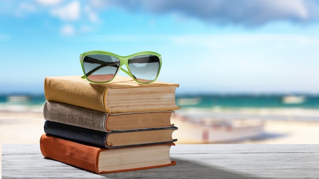 Des livres avec une paire de lunettes de soleil posés sur une table. On voit la plage en arrière plan.