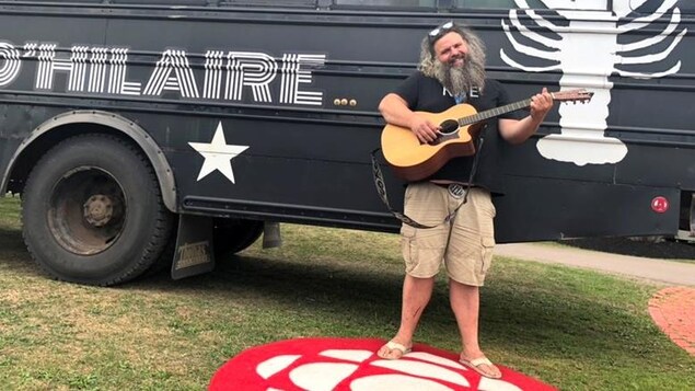 Serge Brideau pose une guitare à la main, devant le bus de son groupe Les Hôtesses d'Hilaire. 
