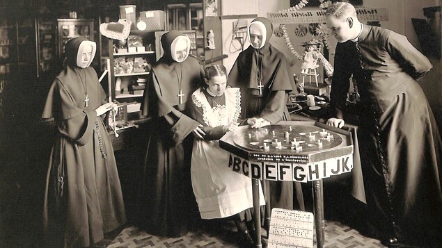 Photo en noir et blanc montrant une jeune fille entourée de trois religieuses et d'un abbé, devant un plateau affichant des lettres de l'alphabet.