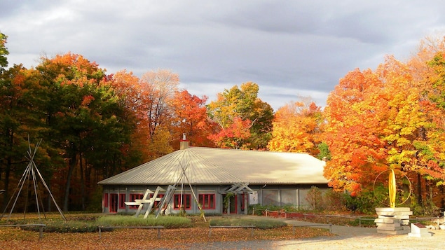 Un bâtiment circulaire devant lequel se trouvent des charpentes de tipis avec un paysage d'automne en arrière-plan.