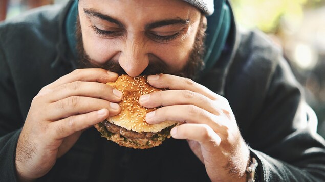 Une personne portant la barbe mange un hamburger avec appétit.