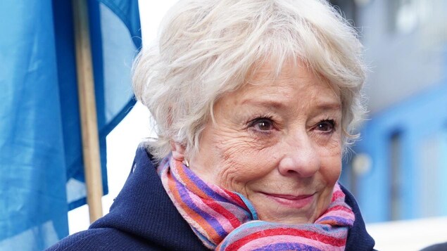 Une femme aux cheveux blanc à l'extérieur. Elle porte un foulard multicolore, un manteau en lainage bleu et un coquelicot.