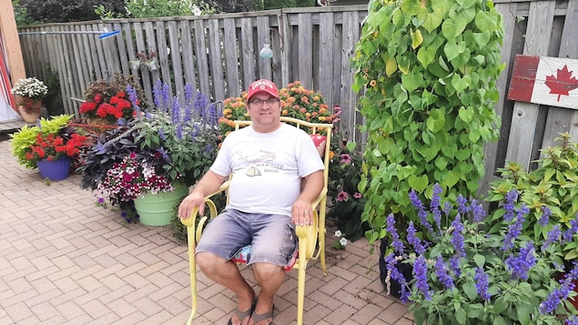 Un homme portant une casquette est assis dans une chaise de parterre dans son jardin et est entouré de plantes et de fleurs.