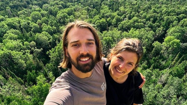 Un homme et une femme se prennent en photo sur une falaise où l'on aperçoit une grande forêt derrière.