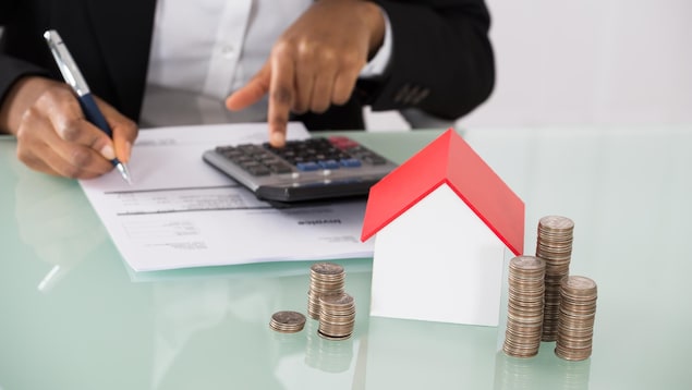 Un homme effectue des calculs et devant lui se tient une petite maison avec des pièces de monnaie.