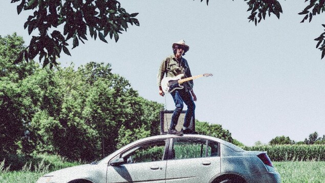 La pochette de l’album Sweet Montérégie d’Alex Burger ; un homme est debout sur le toit d’une voiture avec un chapeau de cowboy, une guitare électrique et un amplificateur