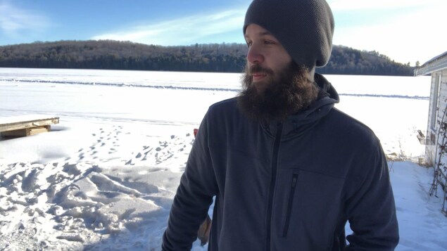 Un homme barbu portant une tuque est debout dehors en plein hiver, sur le bord d’un lac