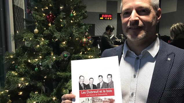 Patrice Dubreuil en studio à Radio-Canada Sudbury, avec une impression de la page couverture « Les Dubreuil et le bois » entre les mains.