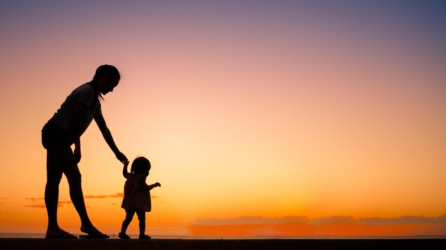 Une silhouette d'une maman et de son jeune enfant. Elle le tient par la mère alors qu'il apprend à marcher. Derrière eux, un coucher de soleil (couleur orange, le soleil n'est plus visible).