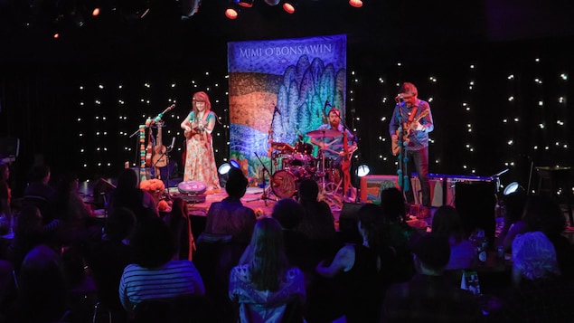 Une photo d'un spectacle de Mimi O'Bonsawin sur scène avec ses musiciens devant un public. C'est écrit «Mimi O'Bonsawin Live in concert».