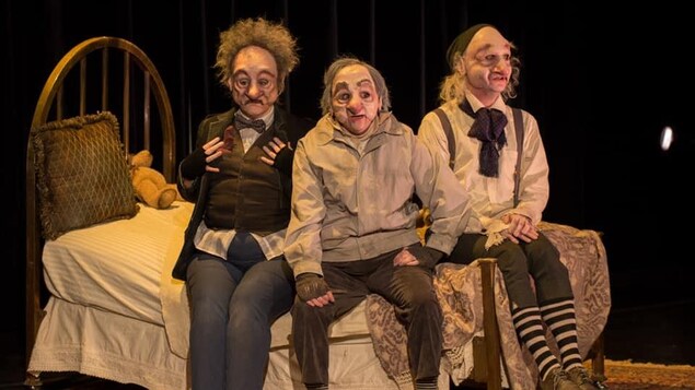 Sur une scène de théâtre, assis sur un lit, trois personnages de vieillards