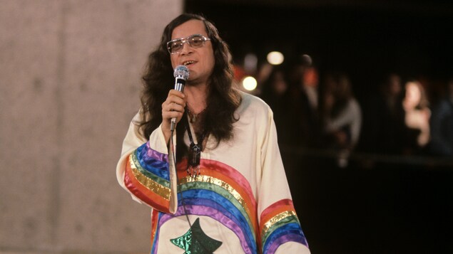 Raoûl Duguay, micro à la main, vêtu d'un poncho orné d'un arc-en-ciel en 1977