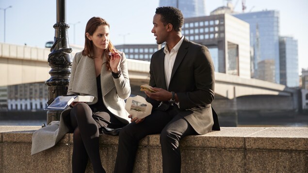 Une femme et un homme en tenue de ville discutent, assis au bord d'un trottoir.