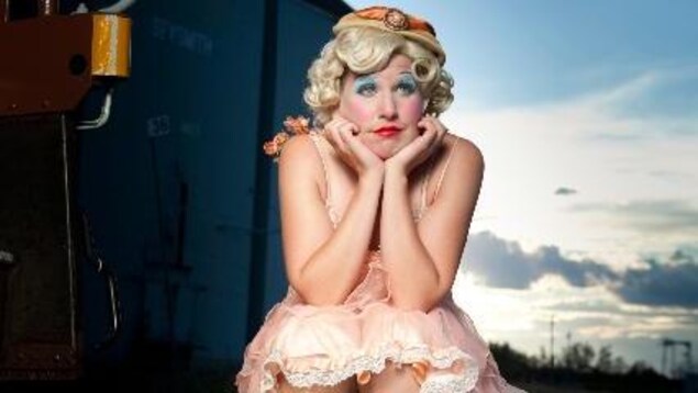 Photographie de la clown Mooky Cornish habillée d'une robe rose pale style années 20. Elle a les paupières bleues et les lèvres rouges. Elle a l’air pensive.