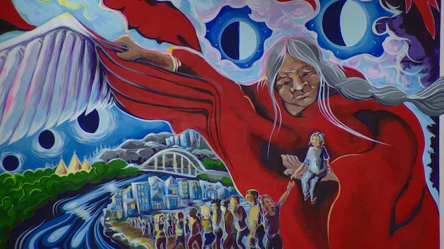 Une murale montre une aînée autochtone qui protège, de son châle transformé en aile, le peuple d'Edmonton qui retourne à la maison.