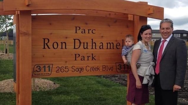 Daniel Vandal et Natalie Duhamel qui porte un enfant au dos devant le panneau d'un parc sur lequel est écrit le nom de son père Ron Duhamel.