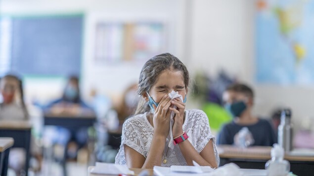 Une élève du primaire abaisse son masque pour se moucher dans un mouchoir. Ses pairs sont assis à leur pupitre individuel en arrière-plan.