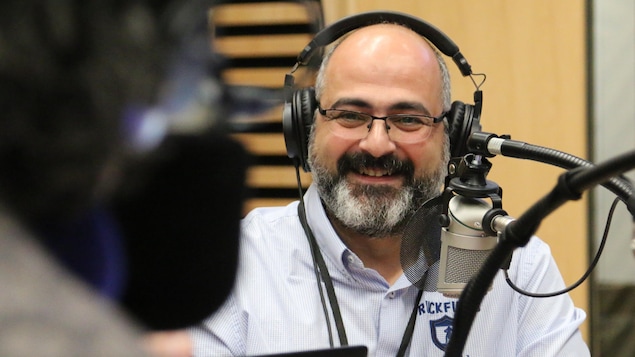 Un homme, casque d'écoute sur la tête, sourit en regardant en direction de l'animateur dans un studio de radio. 