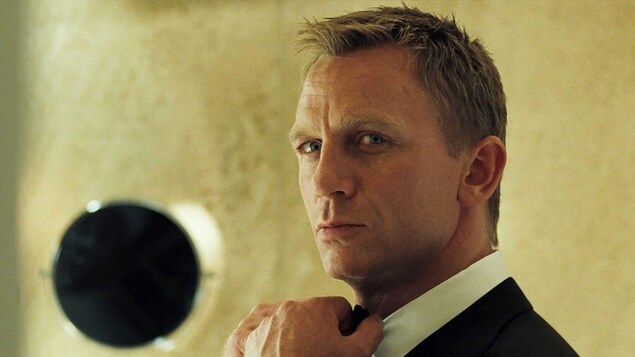 Portrait rapproché de Daniel Craig dans le rôle de James Bond. La tête légèrement tournée, il replace son noeud papillon avec ses mains.
