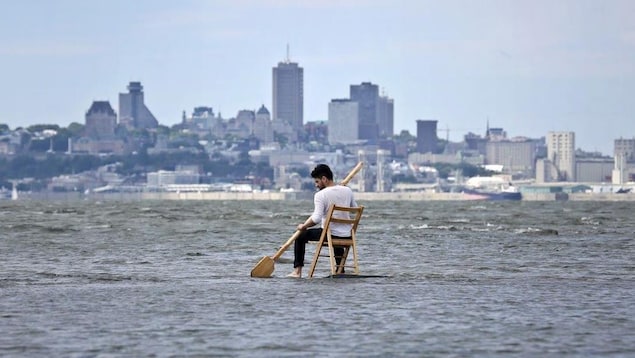 Un homme est assis sur une chaise avec une rame au milieu de l'eau, avec la ville de Québec en arrière-plan. 