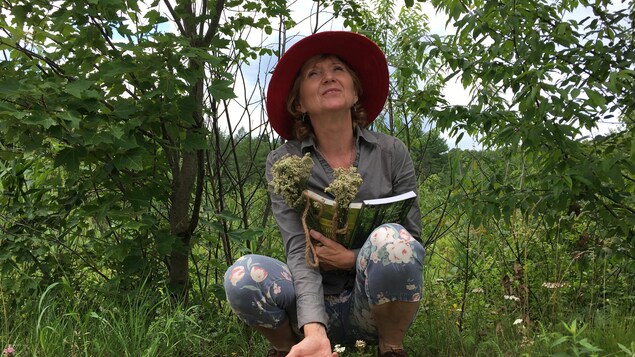 Une femme portant un chapeau accroupie dans un paysage de végétation et tenant des livres et des bouquets de fleurs.