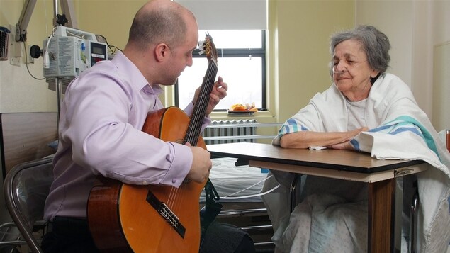 Un homme joue de la guitare dans une chambre d’hôpital pour une patiente.