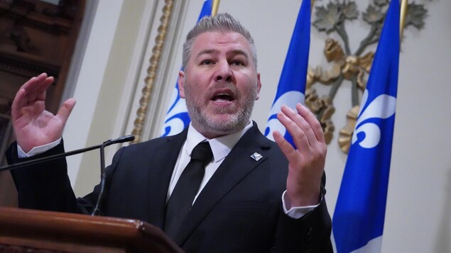 Un homme debout derrière un lutrin et devant des drapeaux du Québec, gesticule en parlant lors d'un point de presse.