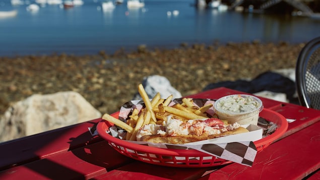 Un plat de frites, salade de choux et d'une guédille est posé sur une table rouge avec des bâteaux sur l'eau au loin.