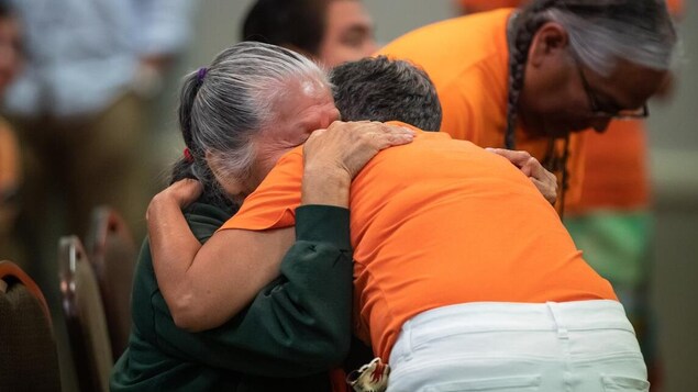 Une personne autochtone âgée pleure dans les bras d'une personne vêtue d'un chandail orange.