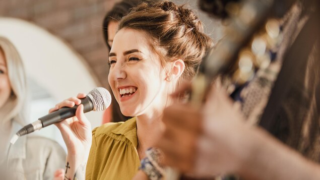 Une femme souriante chante dans un micro entourée d'autres personnes et d'un guitariste.